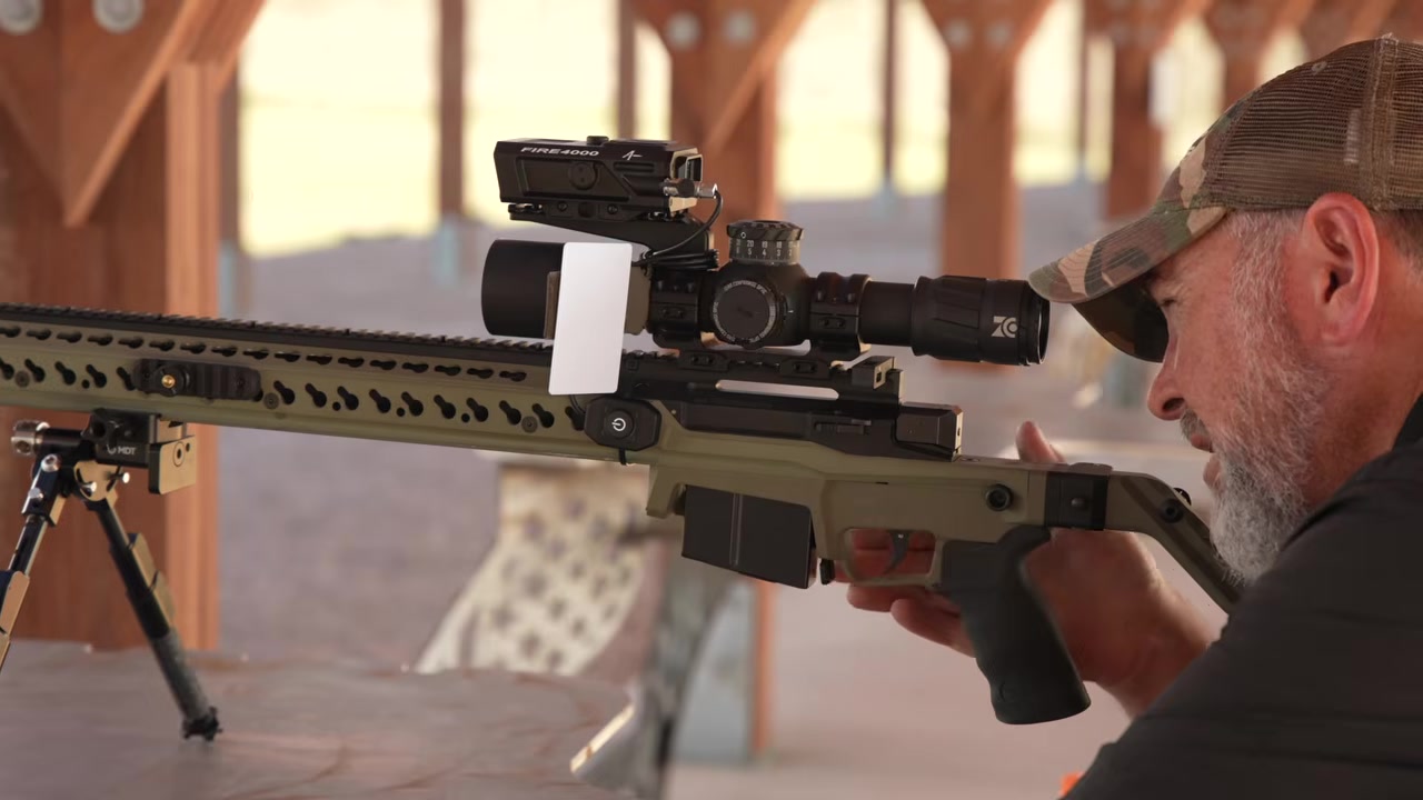 Joel behind his tan chassis rifle at a covered shooting bench, getting ready to confirm his rifle zero, with a scope and a laser rangefinder mounted on top