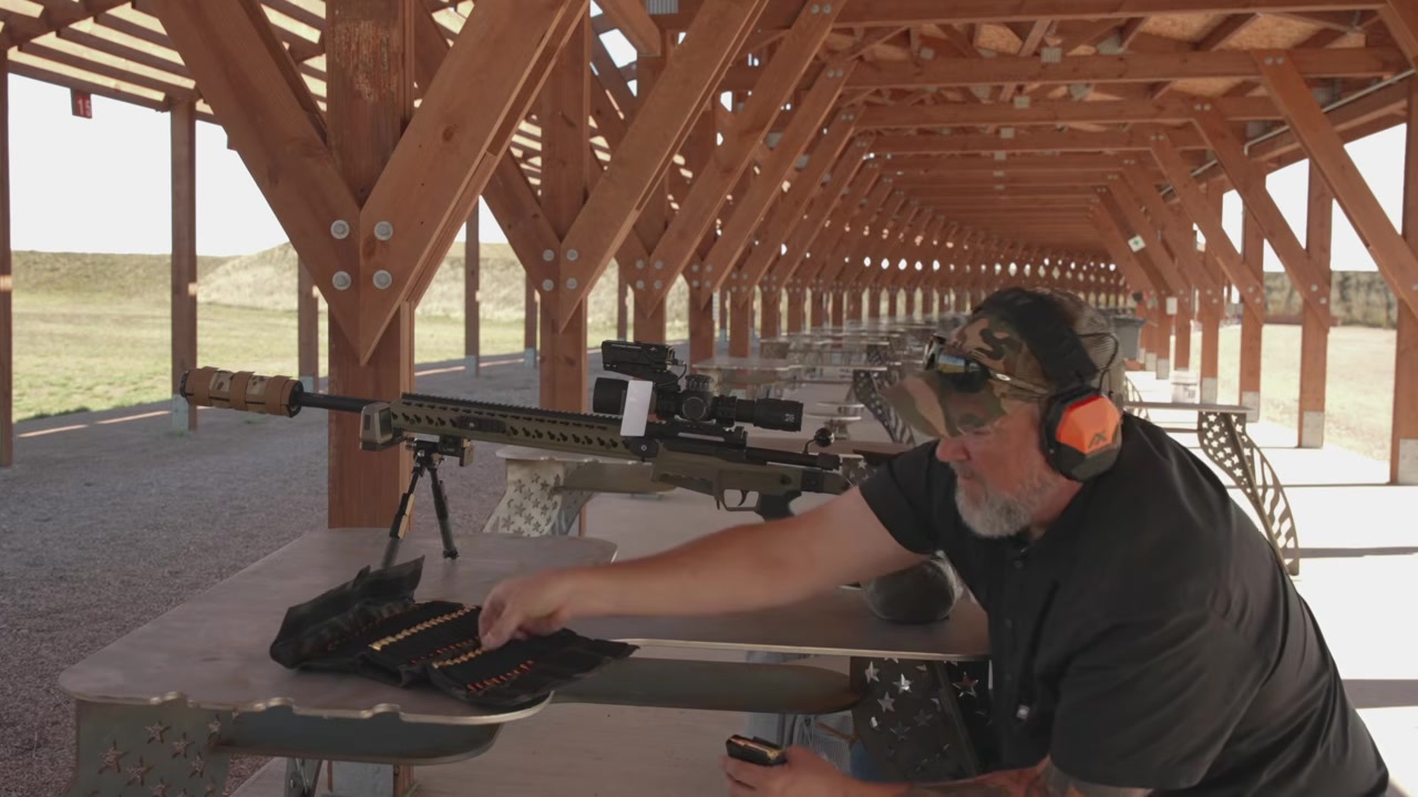 Joel reaching for cartridges in an ammo wallet laid out on the shooting bench next to the rifle
