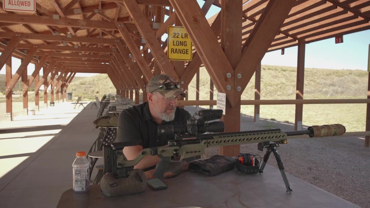 Joel seated behind the rifle on a bipod and rear bag at a level concrete-floored shooting bench